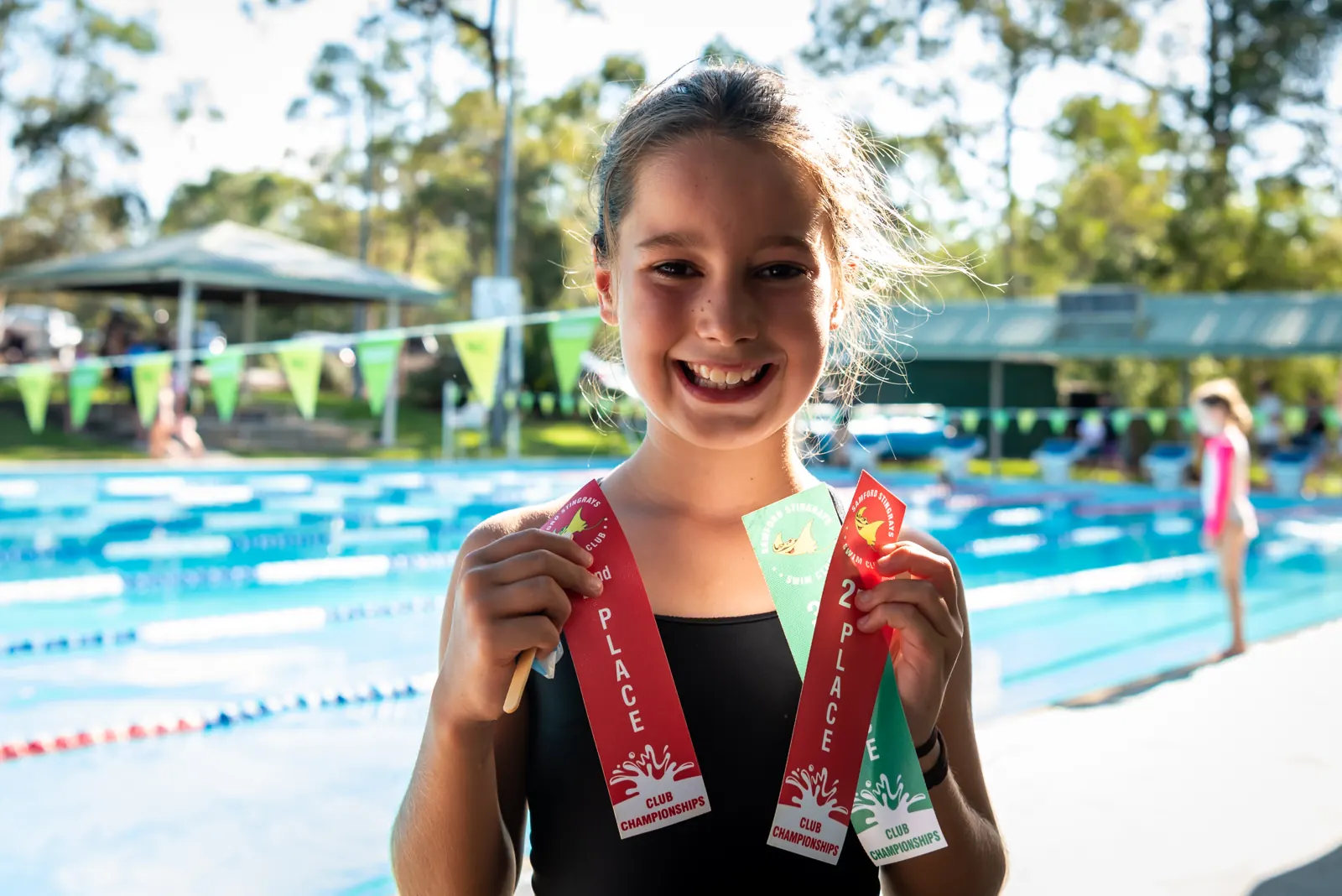 A happy swimmer holding her place ribbons beside the pool